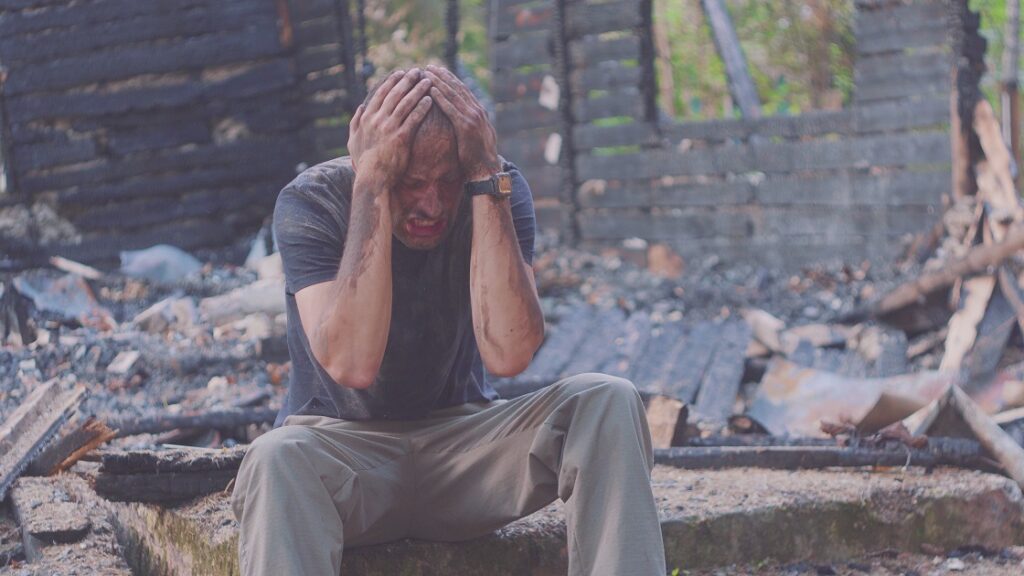 Portrait of a sad man on the background of a burned house, after fire or artillery attack. Consequences of fire disaster accident. Ruins after fire disaster, loss and despair concept